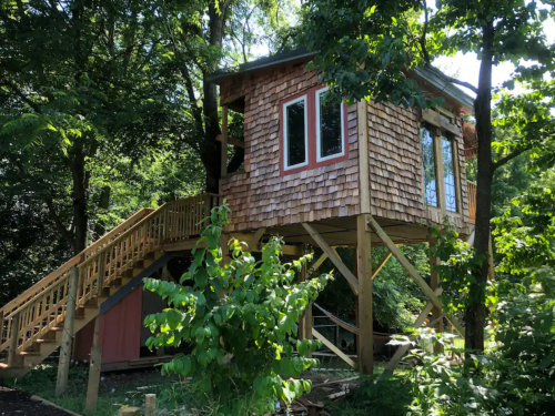 A wooden treehouse with a shingled exterior, elevated on stilts, surrounded by trees and greenery.