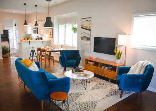 A cozy living room with blue chairs, a coffee table, a TV, and a dining area in the background. Bright and inviting decor.