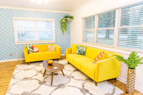 Bright living room with yellow sofas, a round coffee table, patterned walls, and large windows letting in natural light.