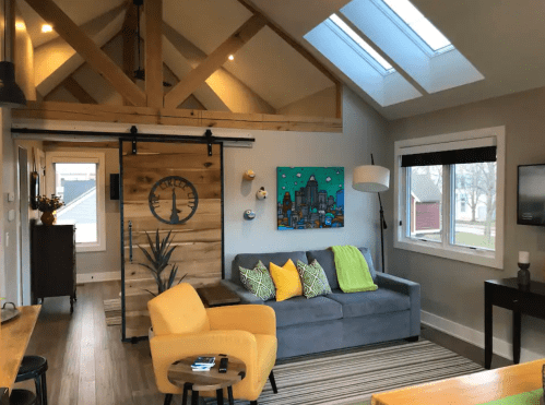 Cozy living room with a gray sofa, colorful pillows, wooden beams, and large windows, featuring a sliding barn door.