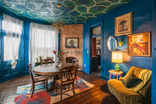 A cozy dining area with a round table, colorful ceiling, blue walls, and vintage decor, featuring a sofa and plants.