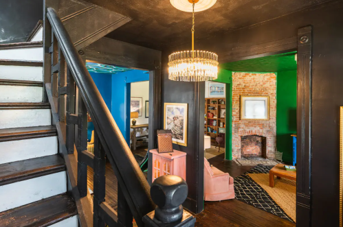 A staircase leads to a colorful living space with a chandelier, exposed brick, and cozy seating areas.