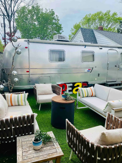 A cozy outdoor seating area with a silver Airstream trailer in the background, surrounded by greenery and decorative elements.
