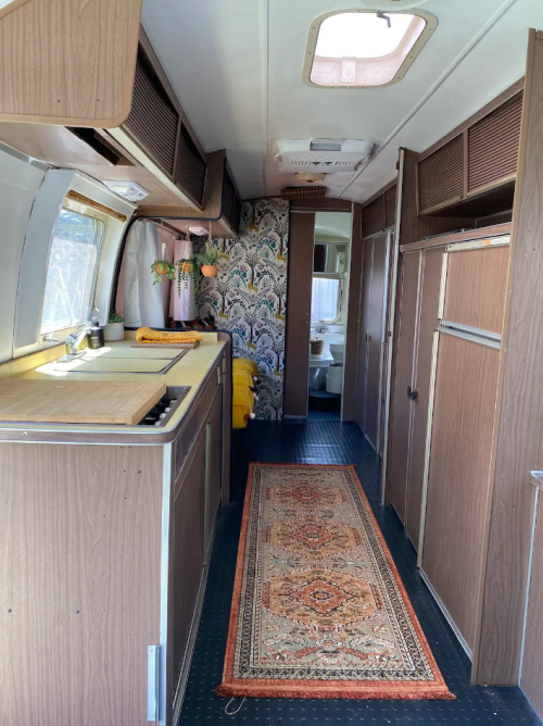 Interior of a vintage trailer featuring a kitchen area, patterned wallpaper, and a decorative rug along the aisle.
