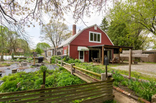 A red barn-style house surrounded by greenery and a garden, with a gravel path leading to the entrance.