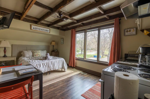 Cozy room with a bed, kitchenette, and large window overlooking a natural view. Warm decor and wooden beams.