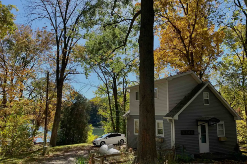 A cozy gray house surrounded by tall trees with autumn leaves, near a serene lake under a clear blue sky.