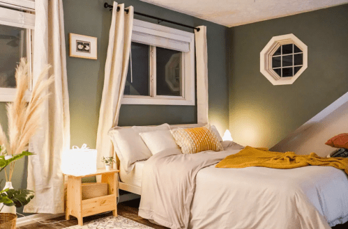 Cozy bedroom with green walls, a bed with yellow accents, and soft lighting from lamps and windows.