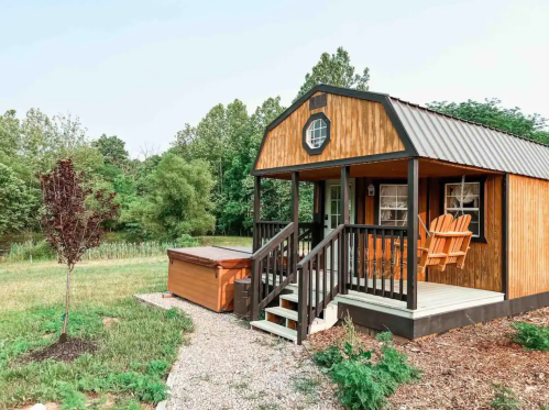 A cozy wooden cabin with a porch and hot tub, surrounded by greenery and a gravel path.