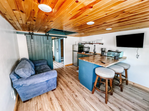 Cozy interior of a small kitchen and living area with a blue couch, bar stools, and wooden ceiling.