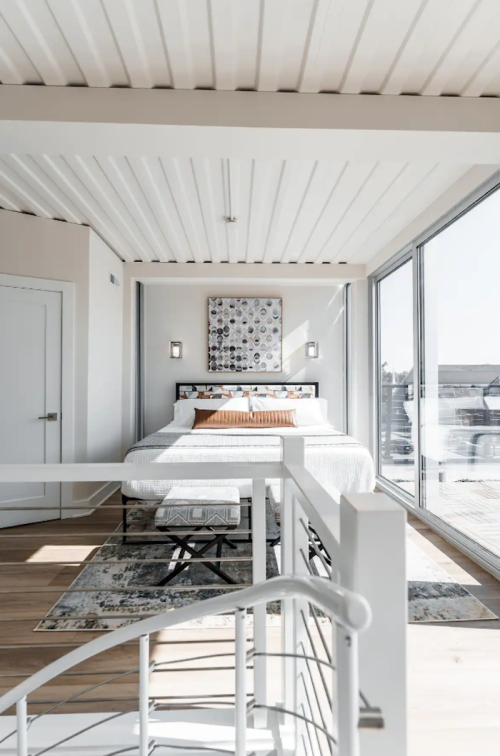 Bright, modern bedroom with large windows, minimalist decor, and a cozy bed featuring decorative pillows.