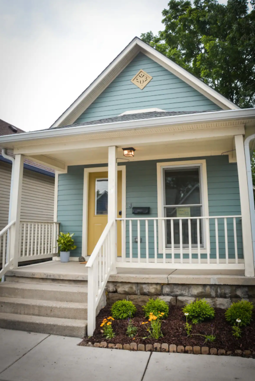 A light blue house with a front porch, white railings, and a small garden.