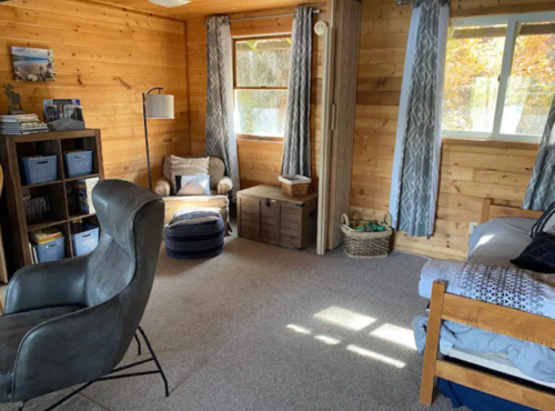 Cozy wooden room with a chair, bed, and natural light from windows, featuring soft furnishings and storage baskets.
