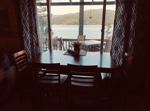 A cozy dining area with a wooden table and chairs, overlooking a scenic lake through large windows.