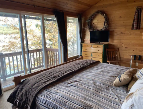 Cozy cabin bedroom with a wooden bed, dresser, TV, and large windows overlooking a scenic view.