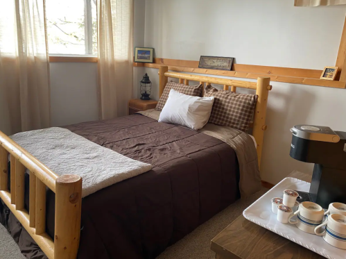 Cozy bedroom with a wooden bed, brown bedding, and a coffee station on a side table. Natural light from a window.