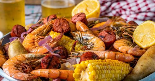 close up of a seafood boil platter with crawfish, shrimp, potatoes, corn on the cob, and lemon wedges next to glasses of beer