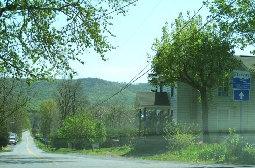 A quiet road lined with trees, leading to a house and a sign for a byway, with mountains in the background.