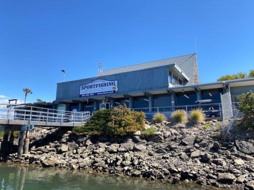 A sportfishing building by the water, featuring a sign and surrounded by rocks and greenery under a clear blue sky.