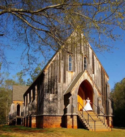 A bride in a white gown stands on the steps of a rustic wooden church surrounded by trees and blue sky.