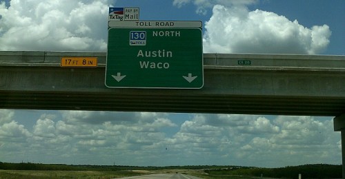 Green highway sign indicating toll road directions to Austin and Waco, with a clear blue sky and clouds above.