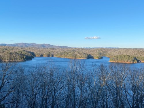 A serene landscape featuring a calm lake surrounded by trees and distant mountains under a clear blue sky.