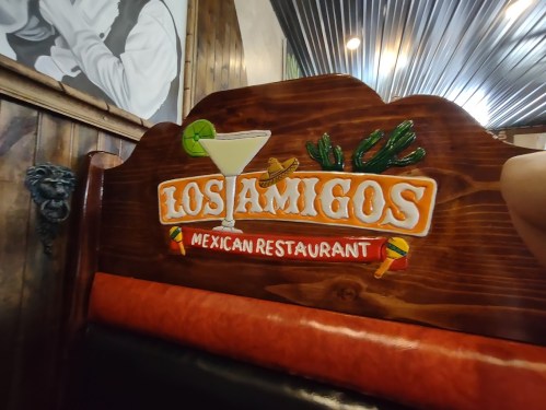 Sign on a wooden restaurant booth reading "Los Amigos Mexican Restaurant" with decorative elements like a margarita and cacti.