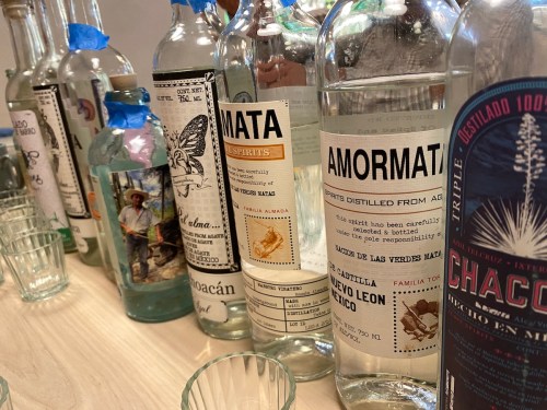 A row of various clear spirit bottles on a table, some with labels and blue tape, alongside small glasses.