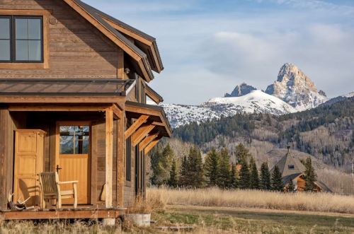 A rustic wooden house with a porch overlooks a snowy mountain landscape under a clear sky.