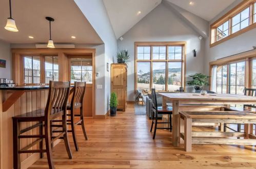 Cozy interior of a modern cabin with wooden floors, a dining table, bar stools, and large windows showcasing mountain views.
