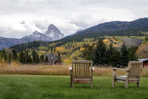 Two wooden chairs sit on a grassy area, overlooking a scenic view of mountains and autumn-colored trees.