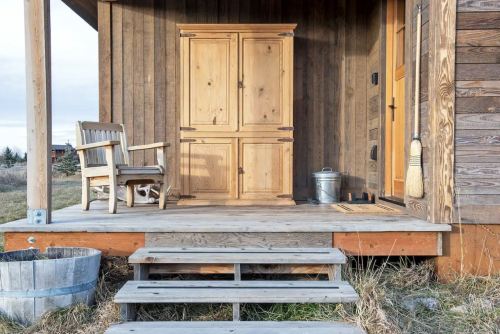 A wooden porch with steps, a chair, a cabinet, and a metal bucket, surrounded by grass and rustic wooden walls.