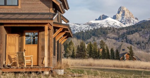 A rustic wooden house with a porch overlooks a mountainous landscape with snow-capped peaks and lush greenery.