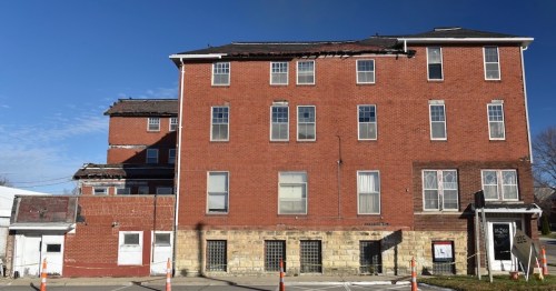 A large, weathered brick building with a damaged roof and multiple windows, set against a clear blue sky.