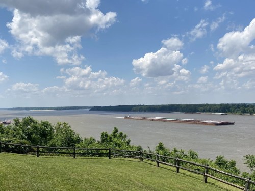 A wide river flows under a blue sky with fluffy clouds, bordered by green trees and a wooden fence in the foreground.