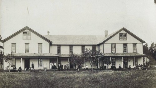 Historic black-and-white photo of a large two-story building with people gathered on the porch and lawn.