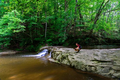 A person sits on a rocky riverbank beside a small waterfall, surrounded by lush green trees.