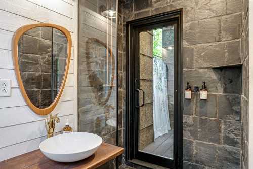 Modern bathroom featuring a round mirror, stone walls, a sleek sink, and a glass door leading to an outdoor area.