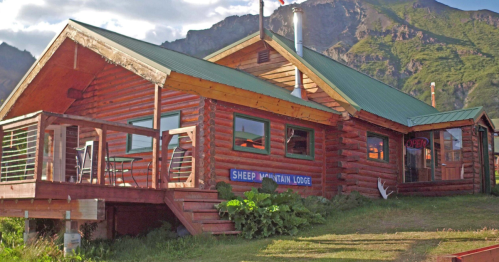 A rustic log cabin with a green roof, surrounded by mountains, featuring a sign that reads "Sheep Mountain Lodge."
