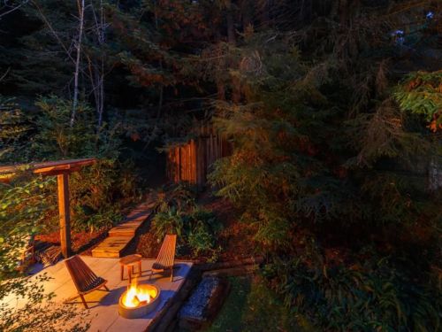 Cozy outdoor space with a fire pit, wooden chairs, and surrounding trees, illuminated at night.