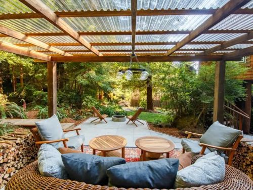 Cozy outdoor seating area with a wooden pergola, surrounded by trees and a fire pit, featuring comfortable chairs and tables.