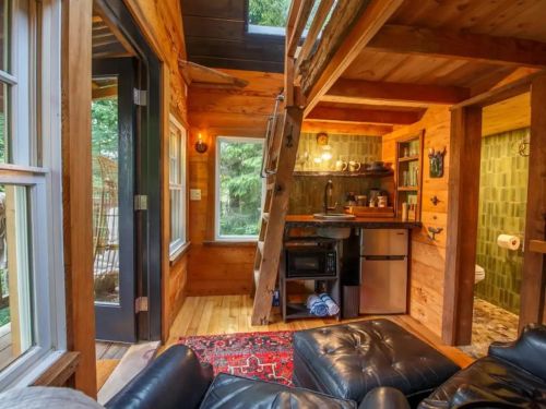 Cozy wooden interior with a loft, kitchenette, and living area featuring a black leather couch and colorful rug.