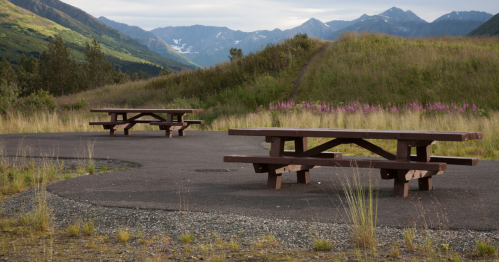 Two wooden picnic tables on a paved area, surrounded by grassy hills and mountains in the background.