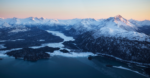 Aerial view of snow-capped mountains and a frozen river, with a serene lake in the foreground during twilight.