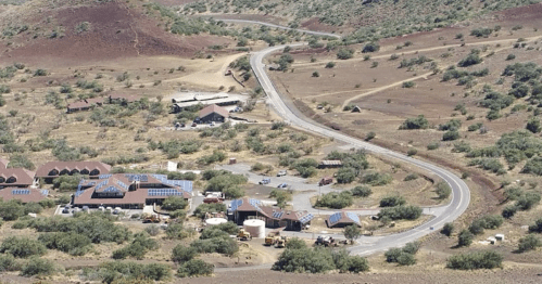 Aerial view of a winding road through a dry landscape with buildings and solar panels in the foreground.