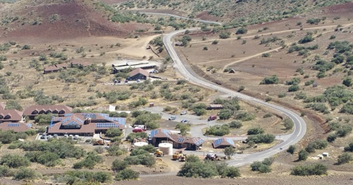 Aerial view of a winding road through a dry landscape with buildings and solar panels in the foreground.