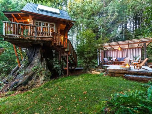 A cozy treehouse with a deck, surrounded by lush greenery and a patio area with seating and a fire pit.