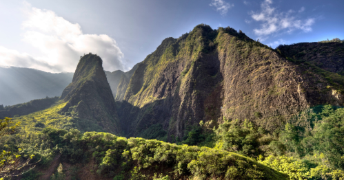 Lush green mountains rise under a blue sky with scattered clouds, showcasing dramatic rock formations and vibrant vegetation.