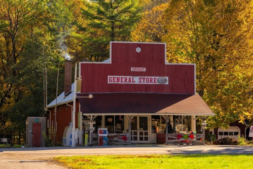 A red general store surrounded by autumn trees, featuring a porch and a chimney with smoke rising.
