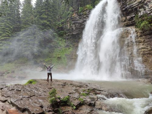 A person stands with arms raised in front of a large waterfall surrounded by lush green trees and mist.